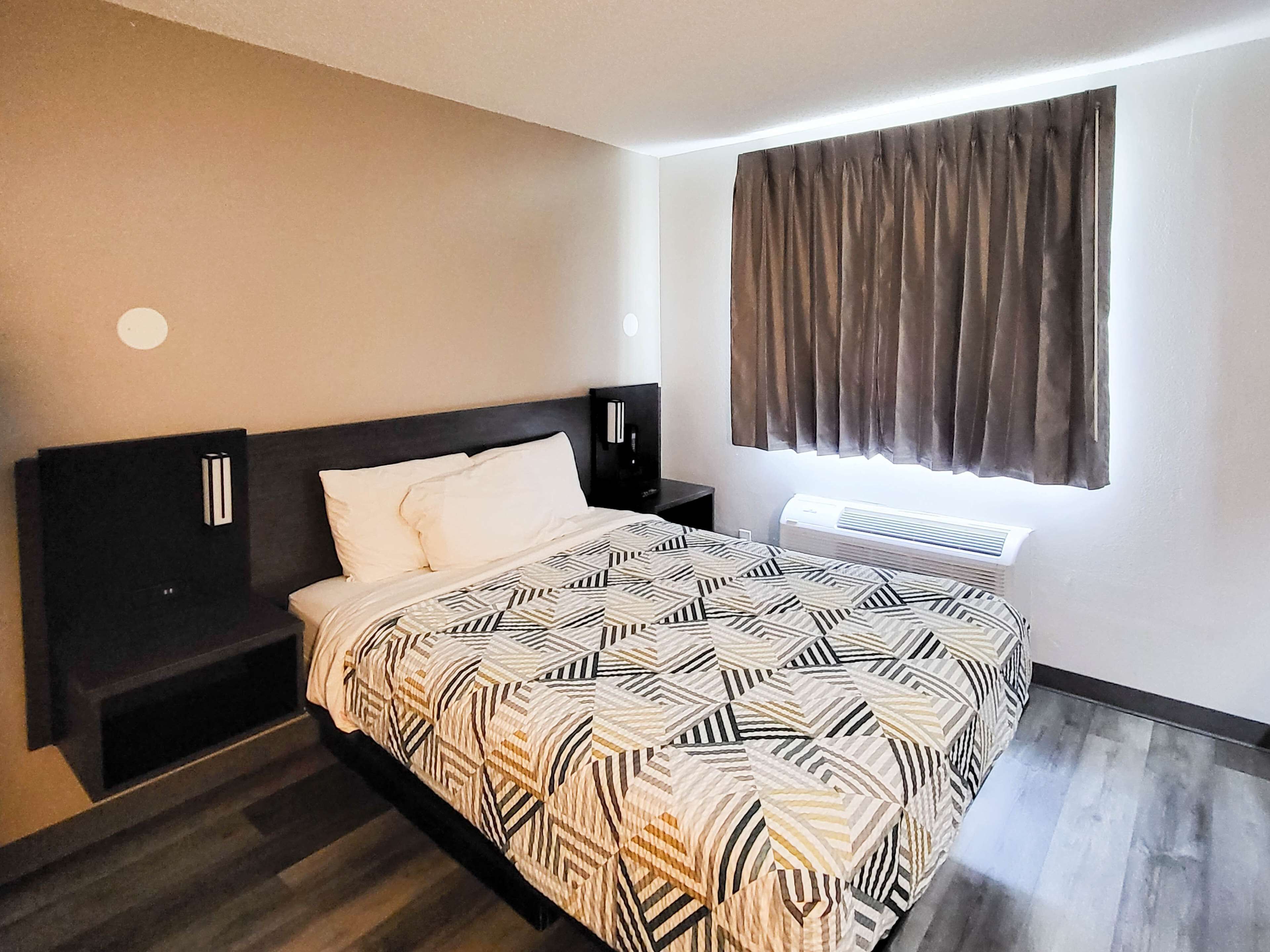 Habitación de hotel con cama, mesita de noche, televisor montado en la pared, ventana con cortinas y aire acondicionado integrado.