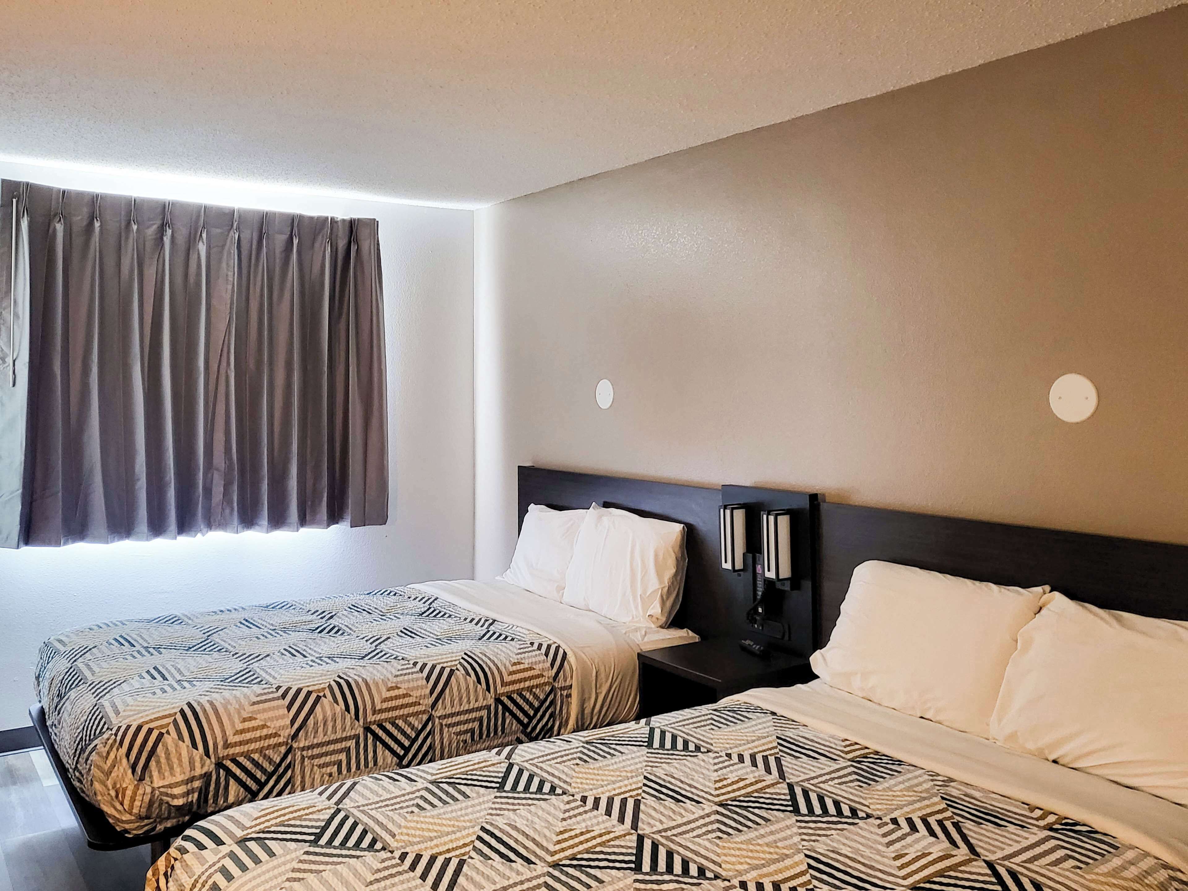 A hotel room with two beds, a window with curtains, a nightstand with a lamp, and a wall-mounted light.
