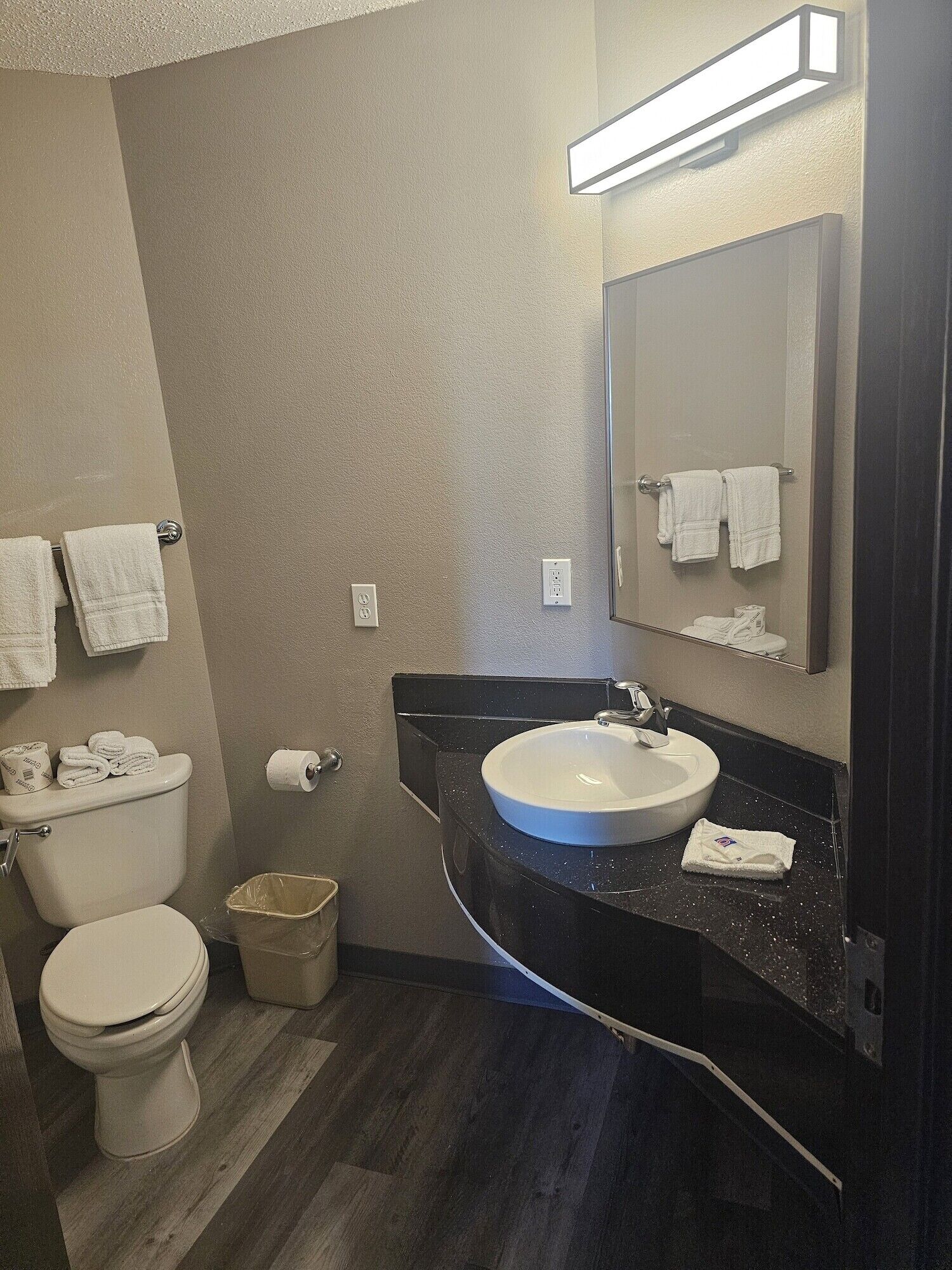 A bathroom with a toilet, sink, and mirror. There are towels hanging on the wall and a trash can.