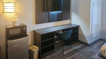 A compact room with a desk, a chair, a microwave, a trash bin, a television mounted on the wall, and a heater.