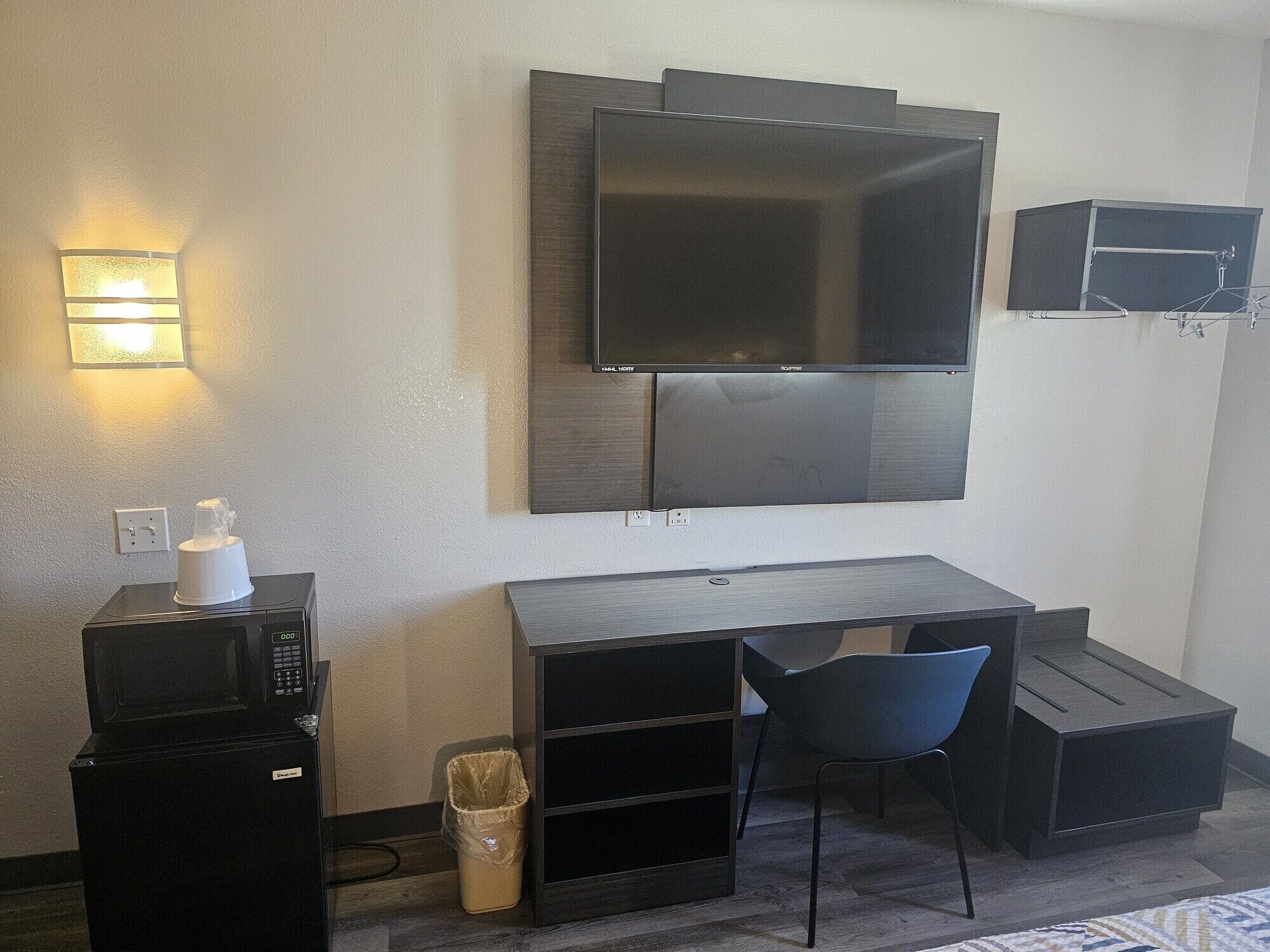 A compact hotel room with a flat-screen TV mounted on the wall, a small desk with a chair, a microwave, and a trash bin.