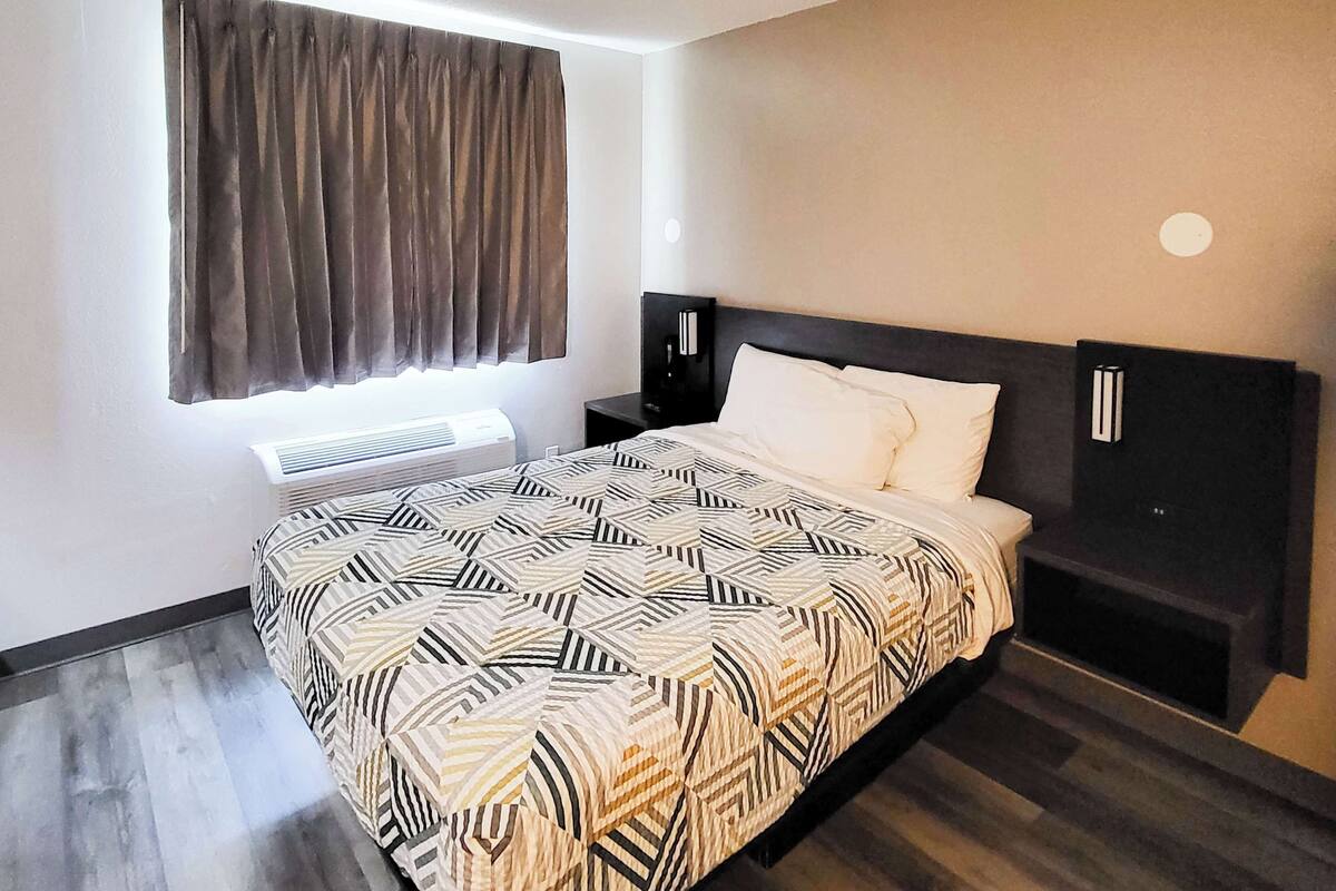 A modern hotel room with a large bed, a bedside table, a wall-mounted TV, and a window with curtains.