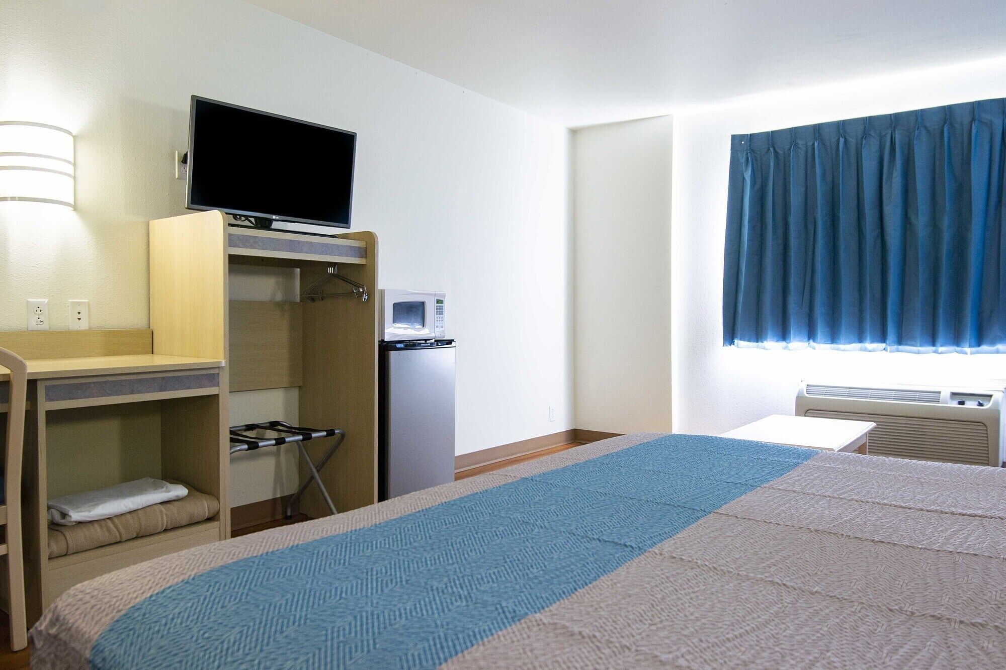 Ein Hotelzimmer mit einem Bett, einem Schreibtisch mit Computer, einer Mikrowelle, einem kleinen Heizgerät und einem Fenster mit Vorhängen.