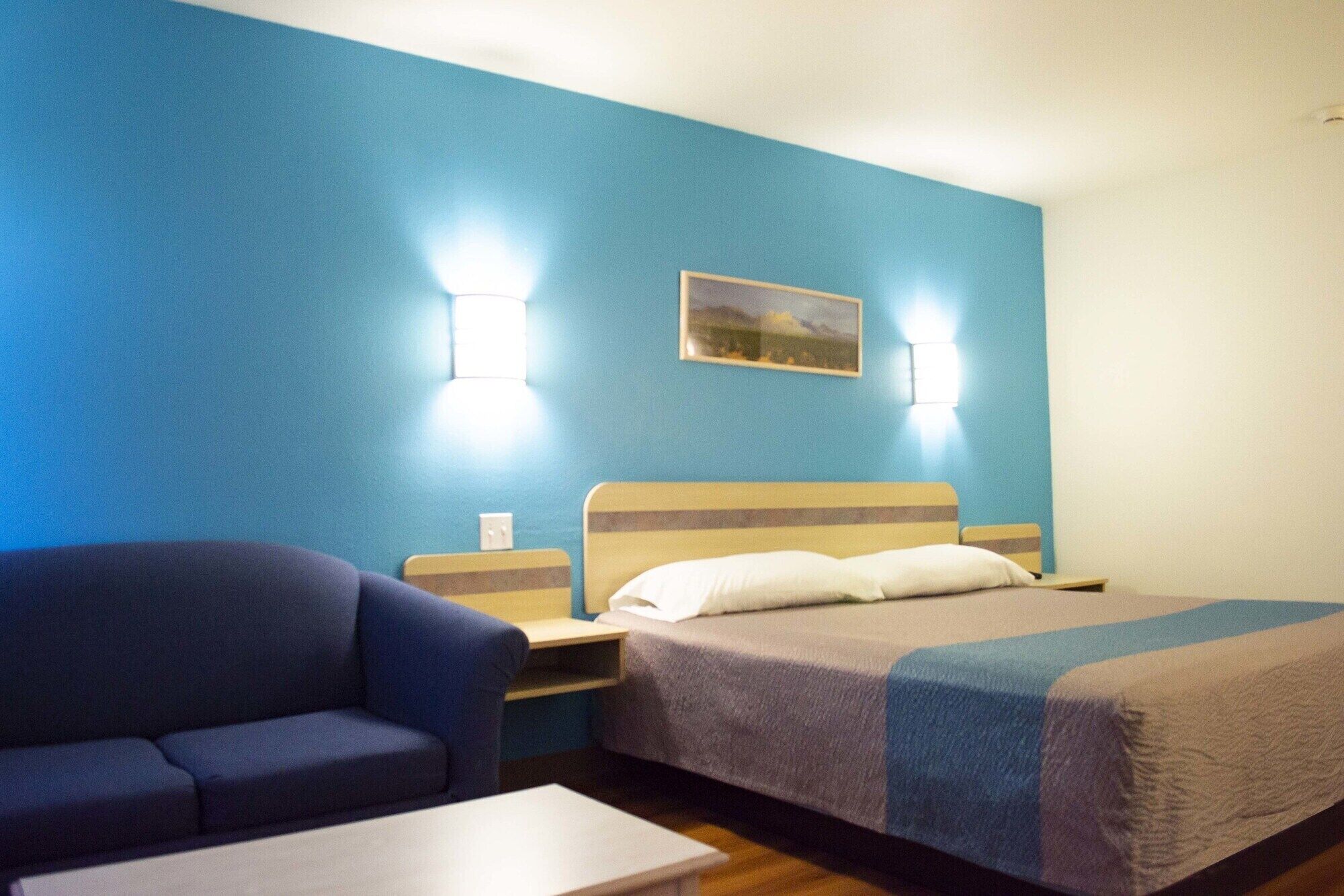 A hotel room with a bed, a sofa, and a small table. There are two wall-mounted lamps and a painting on the wall.