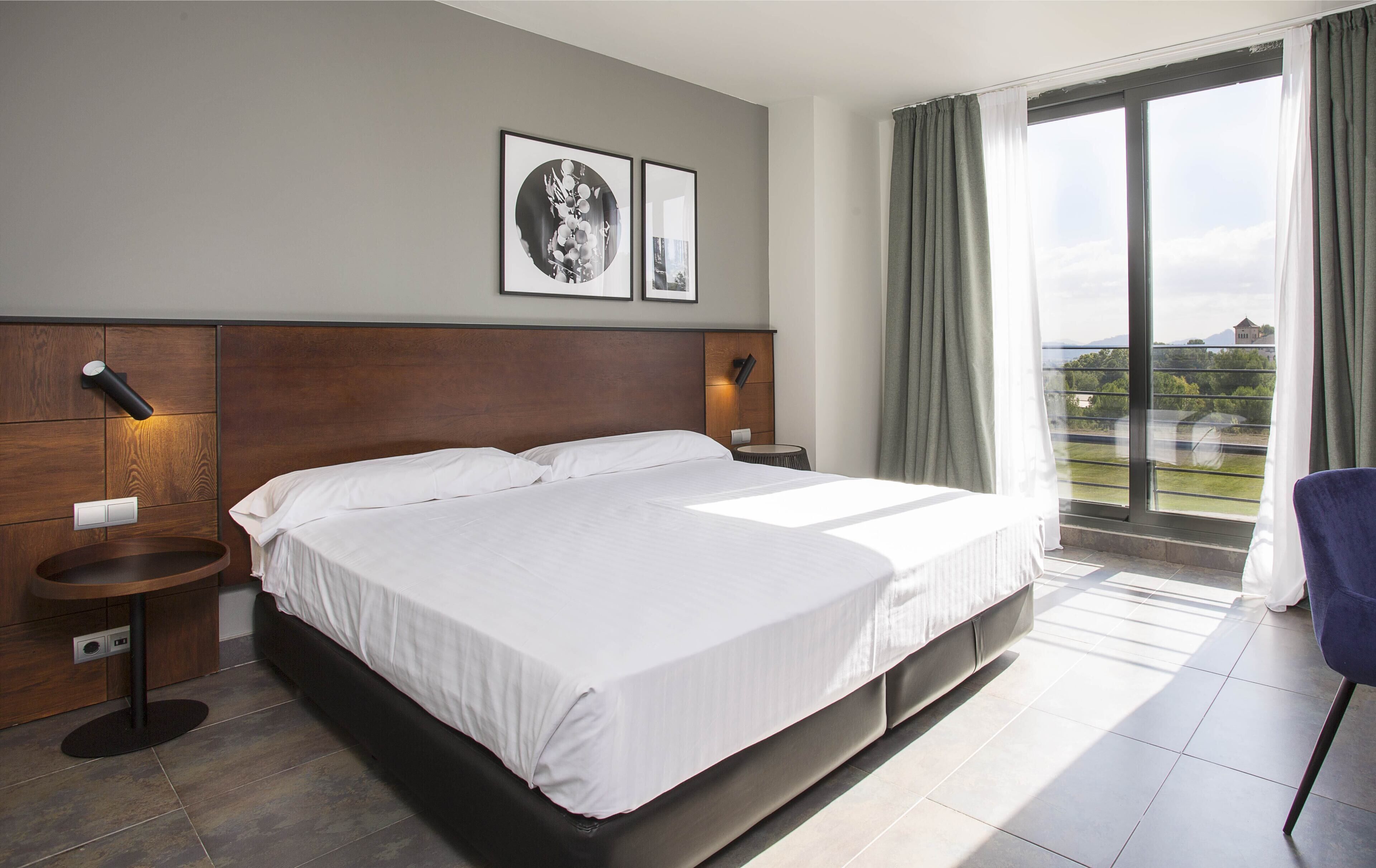 A modern hotel room with a large bed, bedside tables, a round side table, and a view of the outdoors.