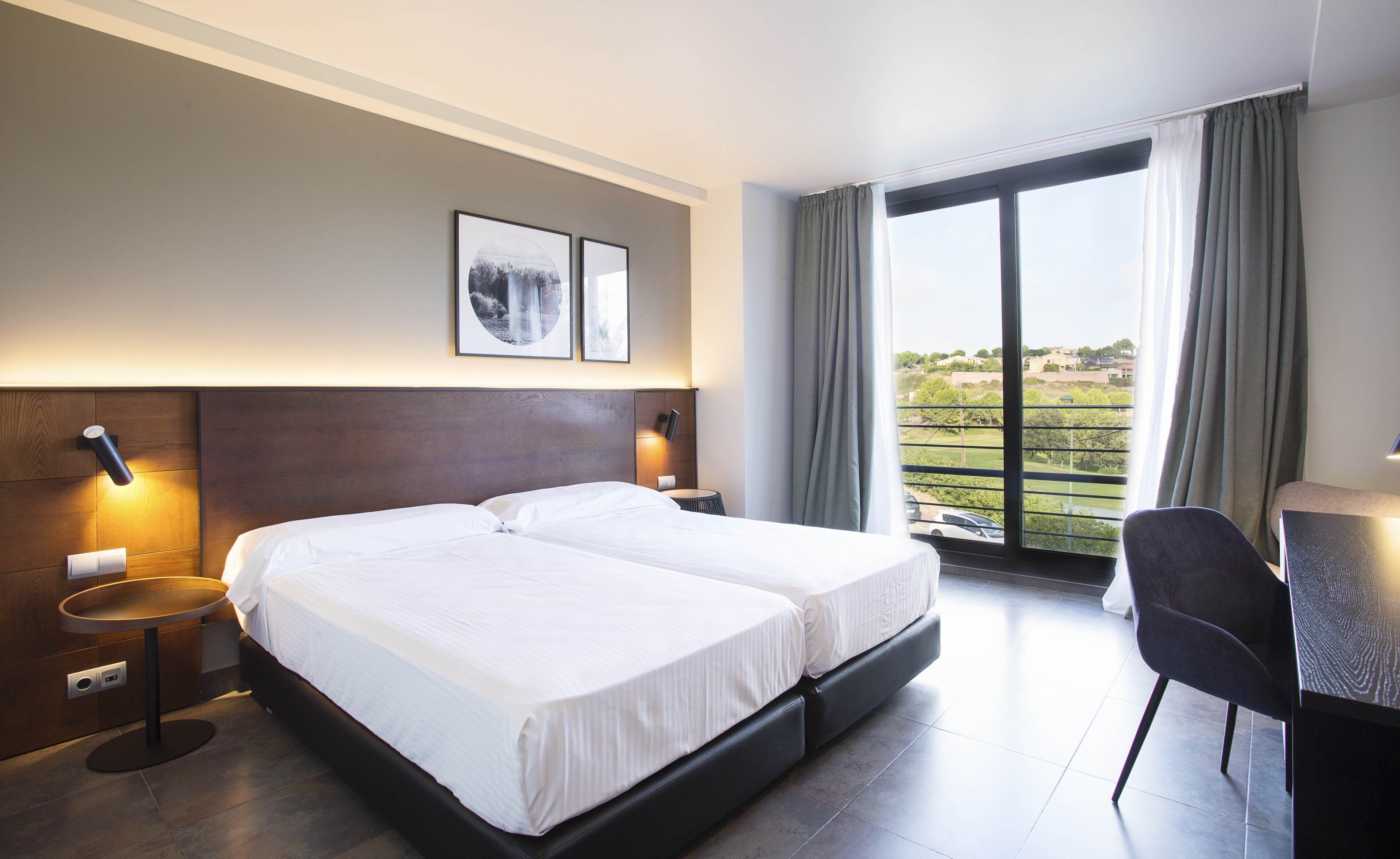 A hotel room with a double bed, bedside tables, a desk, and a large window with curtains.