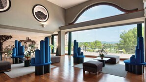 A spacious lobby with modern blue chairs, a wooden floor, and large windows offering a view of greenery and buildings. - Dolce by Wyndham Barcelona Resort (Sant Esteve Sesrovires)