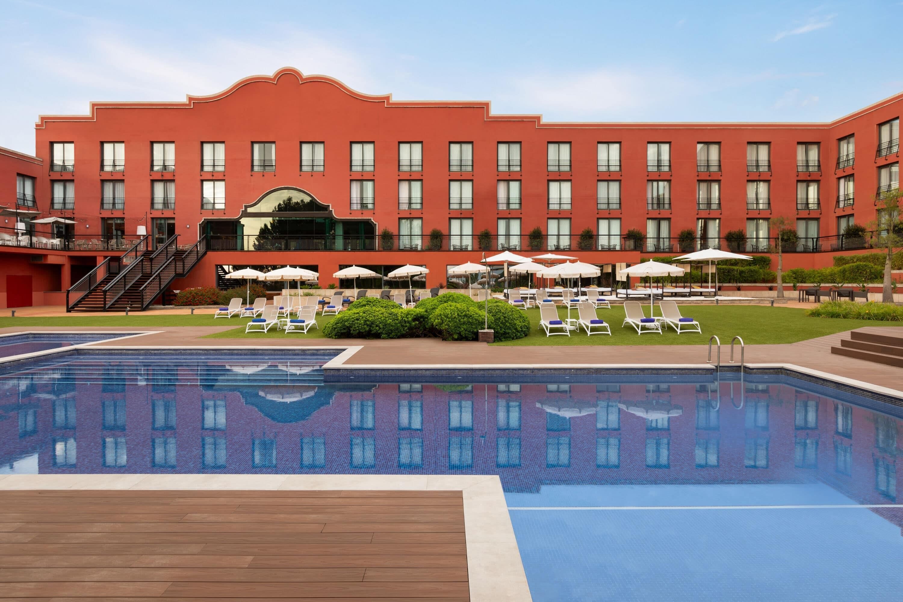 A hotel with a swimming pool, sun loungers, and a large building with multiple windows.
