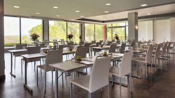 A spacious conference room with multiple tables and chairs, large windows offering a view of greenery, and modern lighting fixtures.