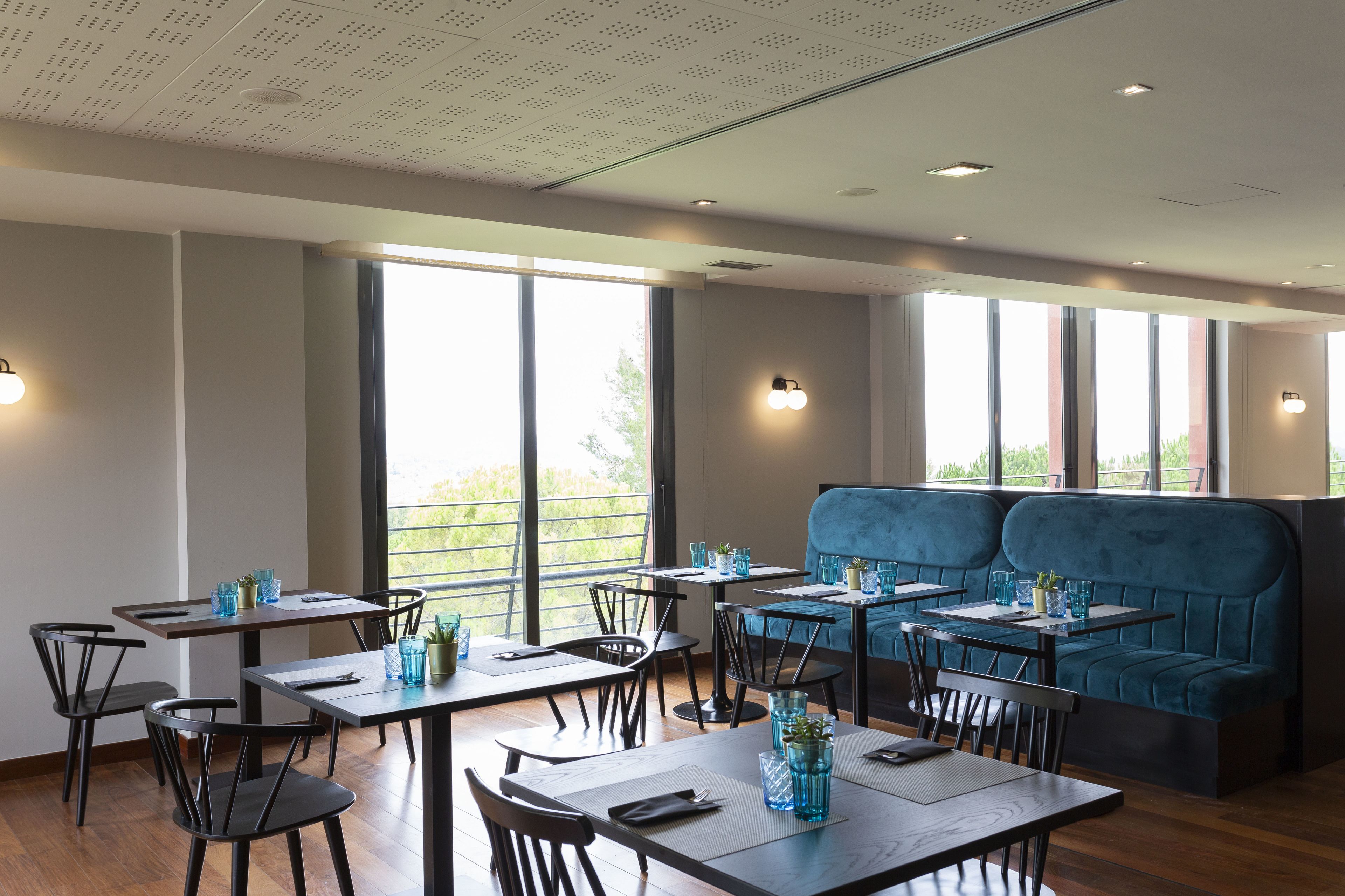 Um restaurante moderno com mesas e cadeiras de madeira, um banco estofado azul e grandes janelas que permitem a entrada de luz natural.