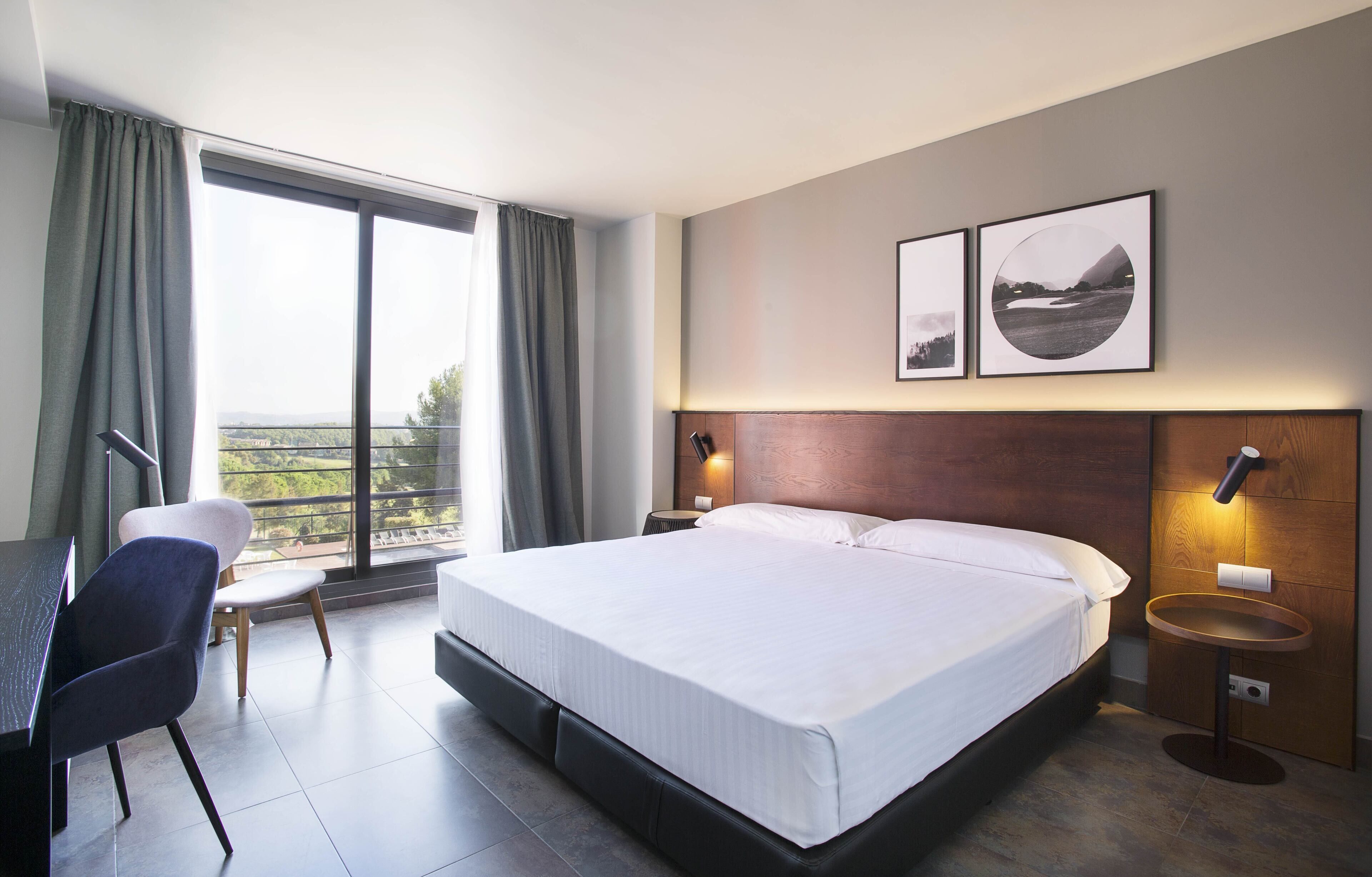 A modern hotel room with a large bed, a desk, and a chair. There are two wall-mounted lamps and a view of the outdoors through a large window.
