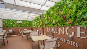 Een zithoek met tafels en stoelen, een houten wand met beplanting, en de tekst 