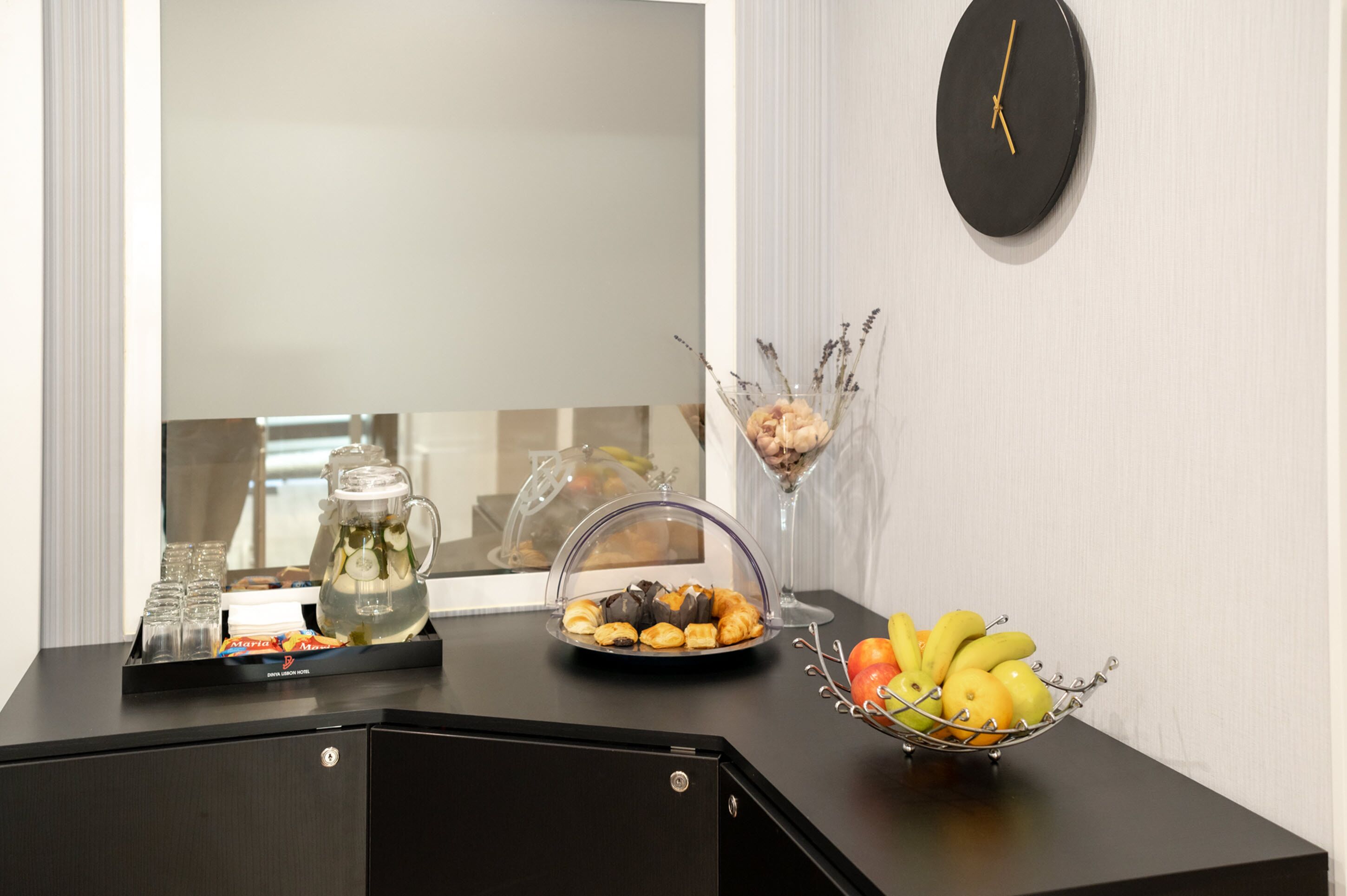 A hotel reception desk with a clock, a fruit bowl, a glass pitcher, and a tray of pastries.