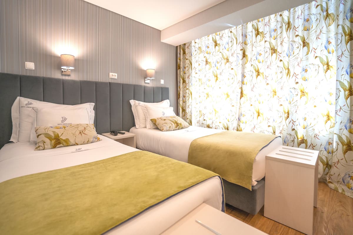 Ein Hotelzimmer mit zwei Betten, einem Nachttisch, einer Lampe und einem Fenster mit floralem Vorhang.