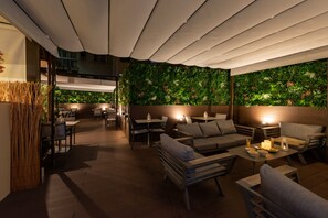Un área de estar al aire libre con mesas y sillas, una pared verde con plantas y un techo de madera.