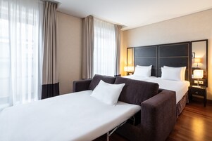 Una habitación de hotel con cama, dos mesitas de noche, un sofá y un ventanal con cortinas.