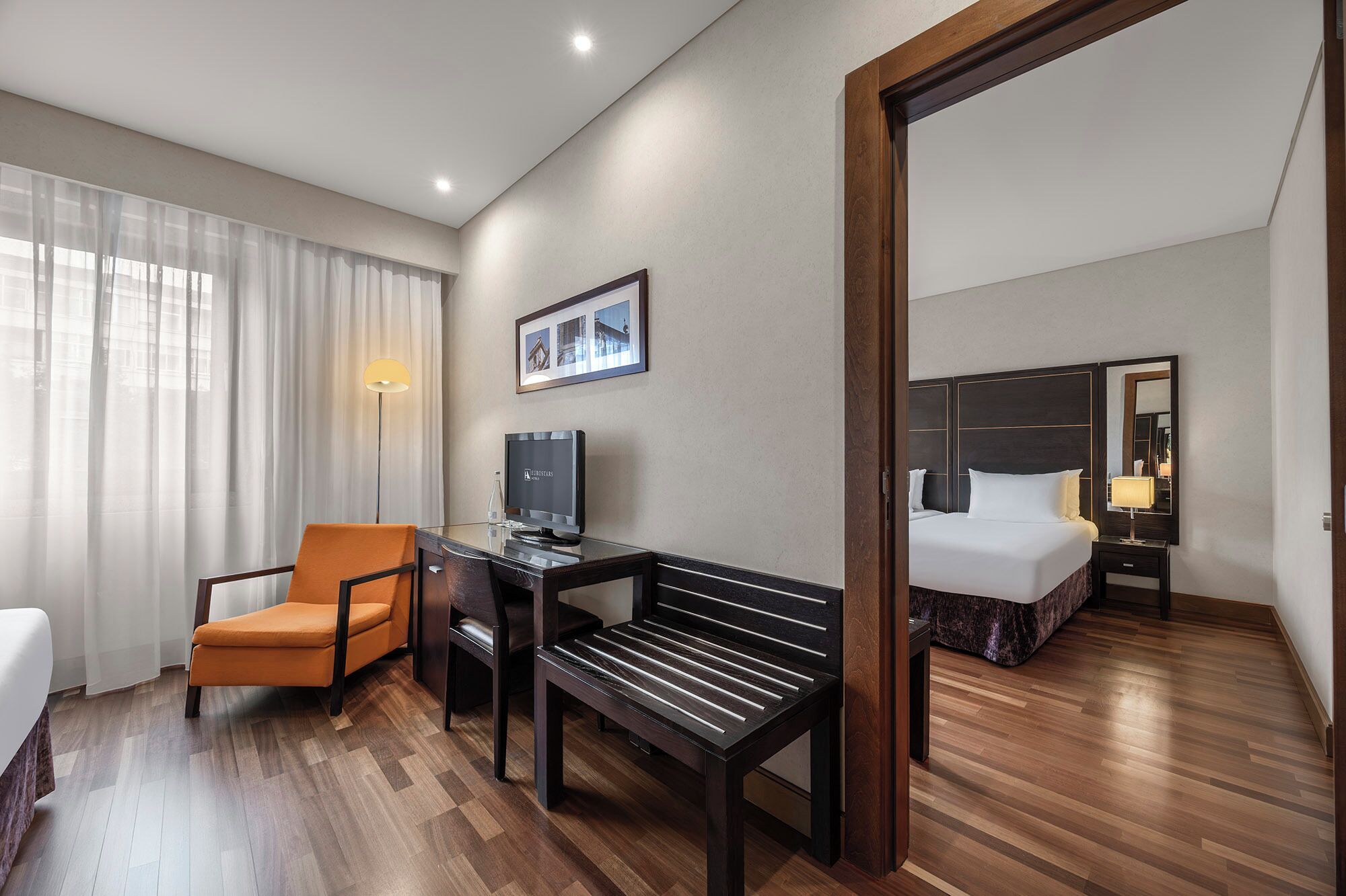A hotel room with a bed, a desk with a computer, a chair, a bench, and a large mirror.