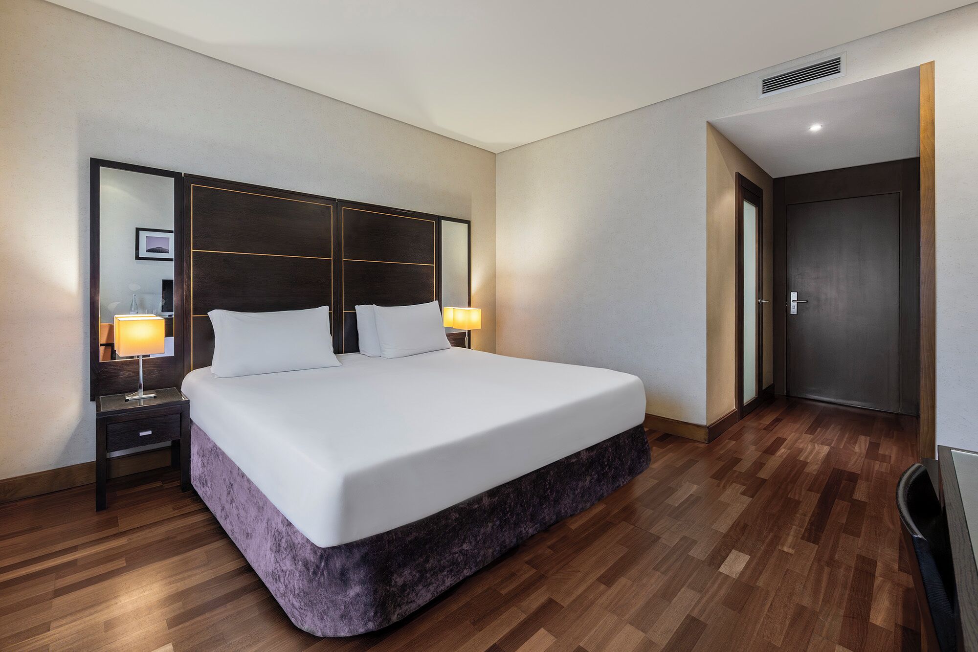 A modern hotel room with a large bed, a nightstand with a lamp, a mirror, and a door to the right.