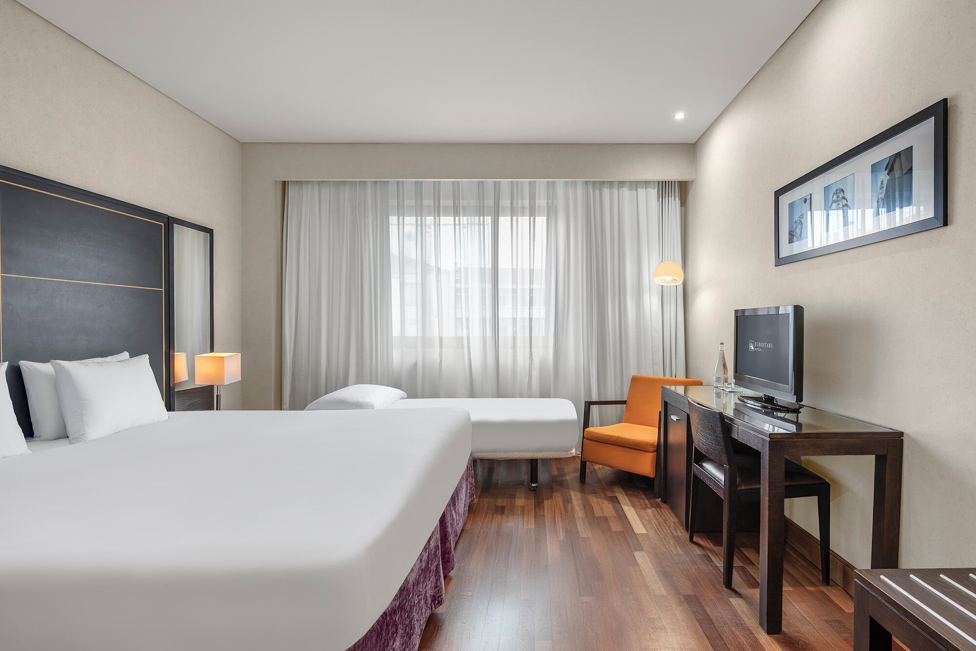 A hotel room with a large bed, a desk with a television, a chair, and a window with sheer curtains.