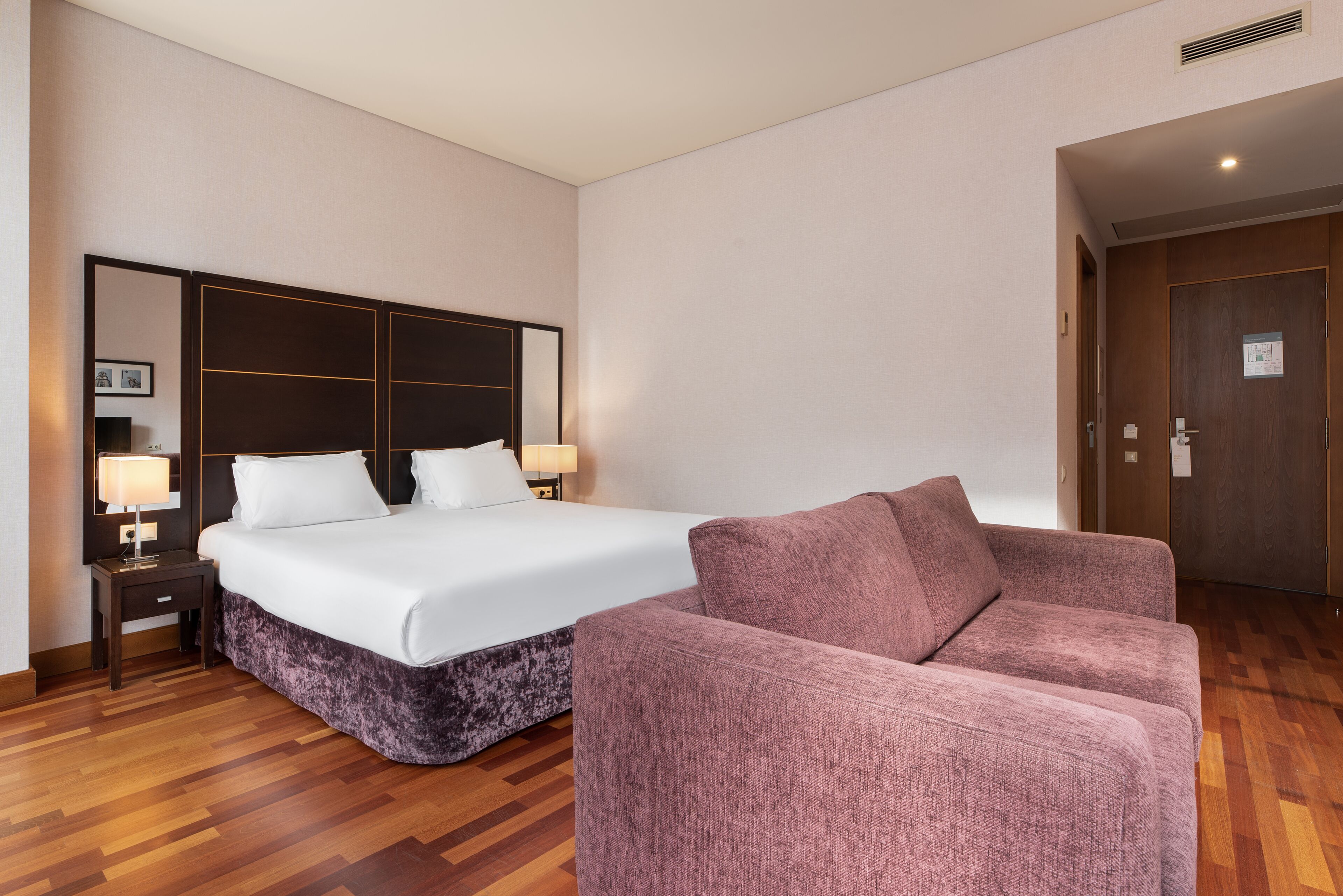 Habitació d'hotel amb un llit gran, una tauleta de nit amb una làmpada, un butac armchair rosa i un terra de fusta.