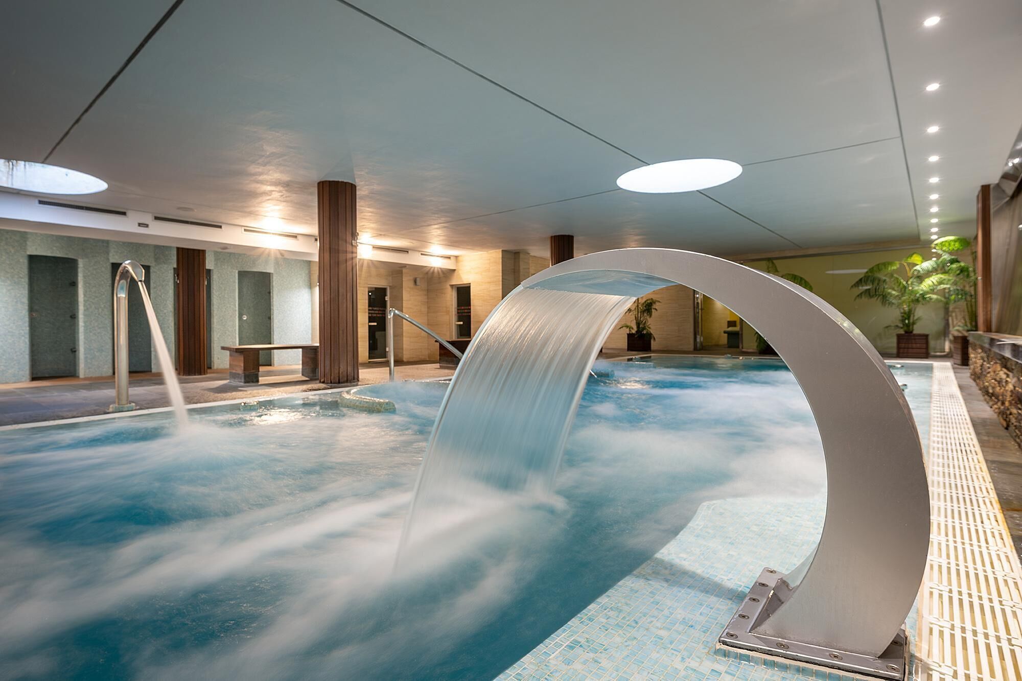 An indoor swimming pool with a waterfall feature, a bench, and a potted plant.