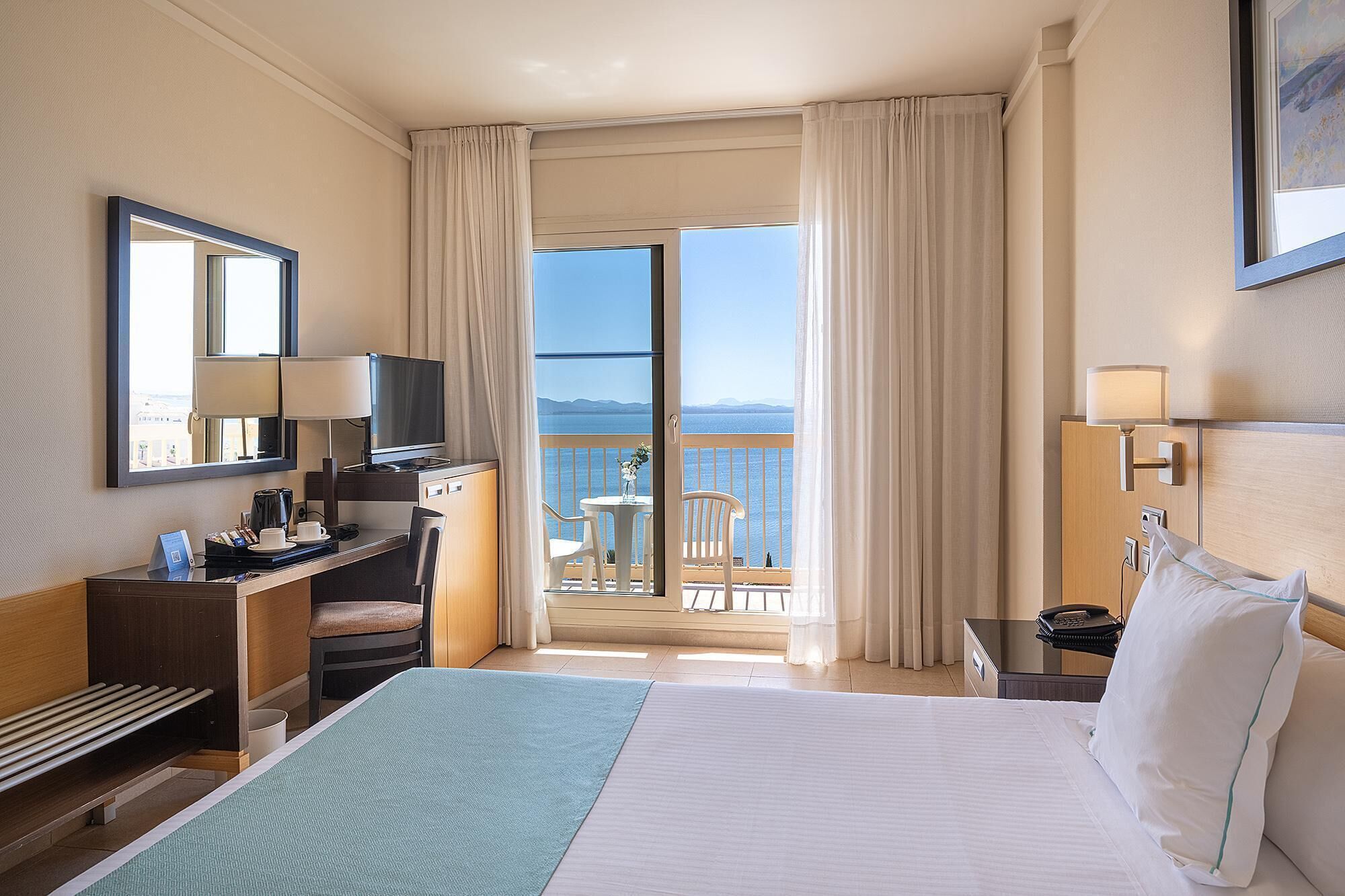 A hotel room with a bed, desk, chair, and a balcony with a view of the sea.