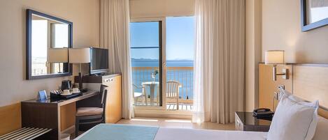 A hotel room with a bed, desk, chair, and a balcony with a view of the sea.