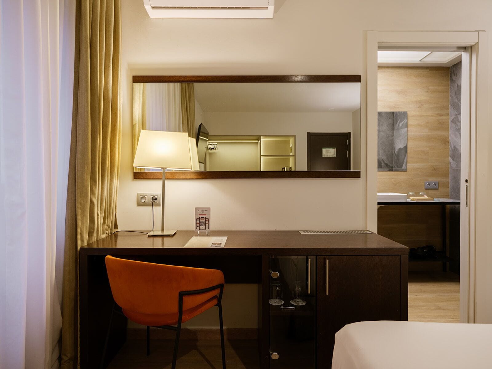 A hotel room with a desk, chair, bed, and a mirror reflecting a bathroom.