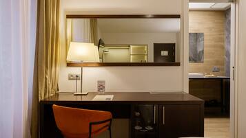 A hotel room with a desk, chair, bed, and a mirror reflecting a bathroom.