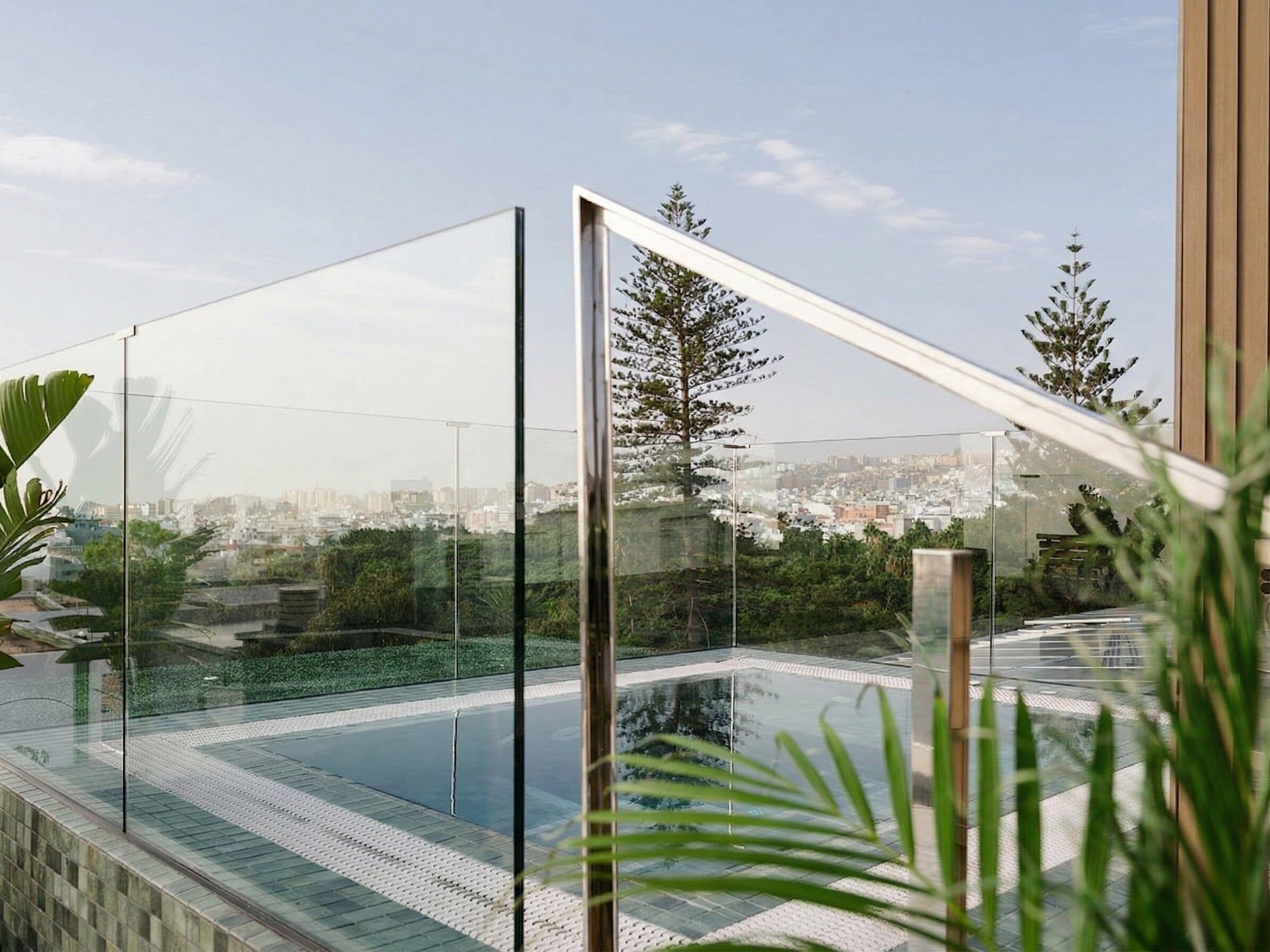 A modern outdoor pool area with glass railings, a clear view of the cityscape, and lush greenery.