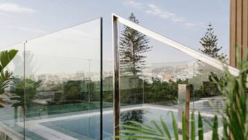 A modern outdoor pool area with glass railings, a clear view of the cityscape, and lush greenery.