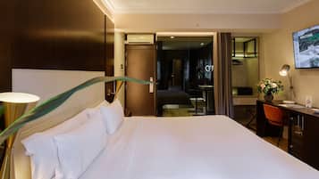A hotel room with a large bed, a desk with a chair, a desk lamp, a vase with flowers, and a television on the wall.