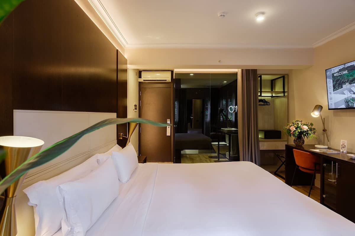 Una habitación de hotel con una cama grande, un escritorio con una silla, una lámpara de escritorio, un jarrón con flores y un televisor en la pared.