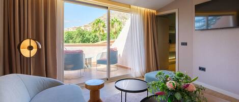 A modern hotel room with a curved sofa, a small round table with a vase of flowers, a floor lamp, and a large sliding glass door leading to an outdoor seating area with a view of greenery and hills.