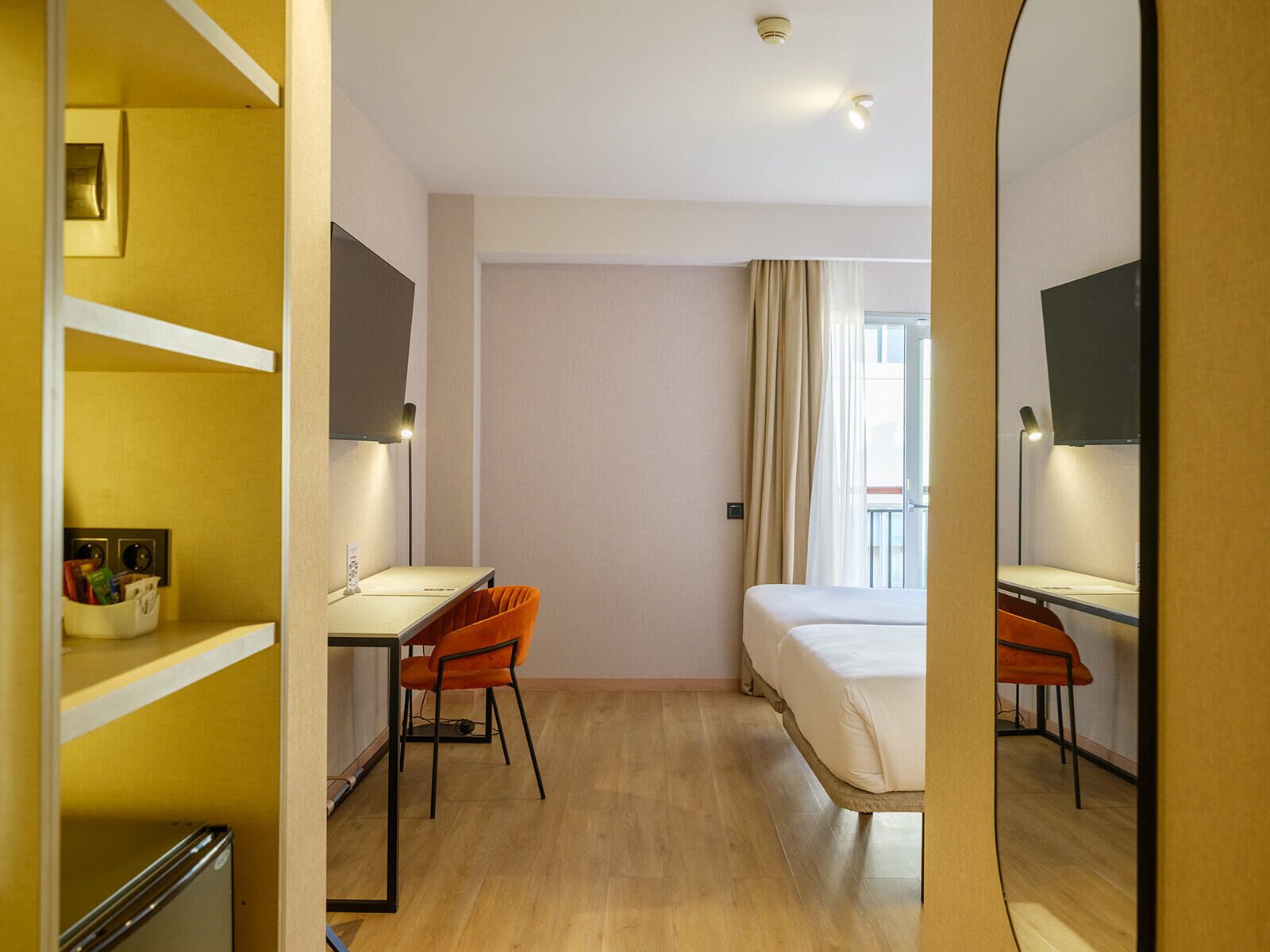 A hotel room with a bed, two desks, a mirror, and a window with curtains.