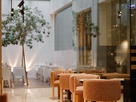 Interior moderno de restaurante com área de estar, mesas e cadeiras de madeira, divisória de vidro e uma janela grande.