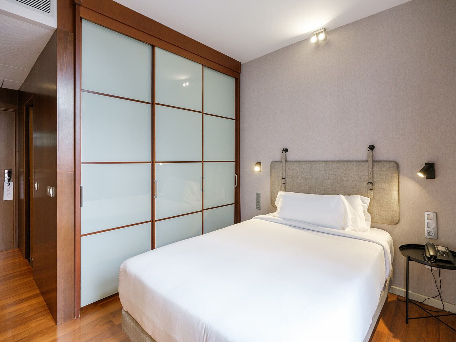 Hotelkamer met een bed, nachtkastje, wandlamp en een schuifdeur met geëtste glaspanelen.
