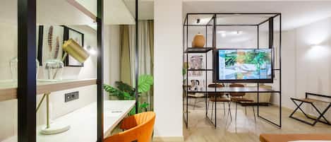 A modern hotel room with a wooden floor, a desk with a chair, a TV, a bed, and a plant.