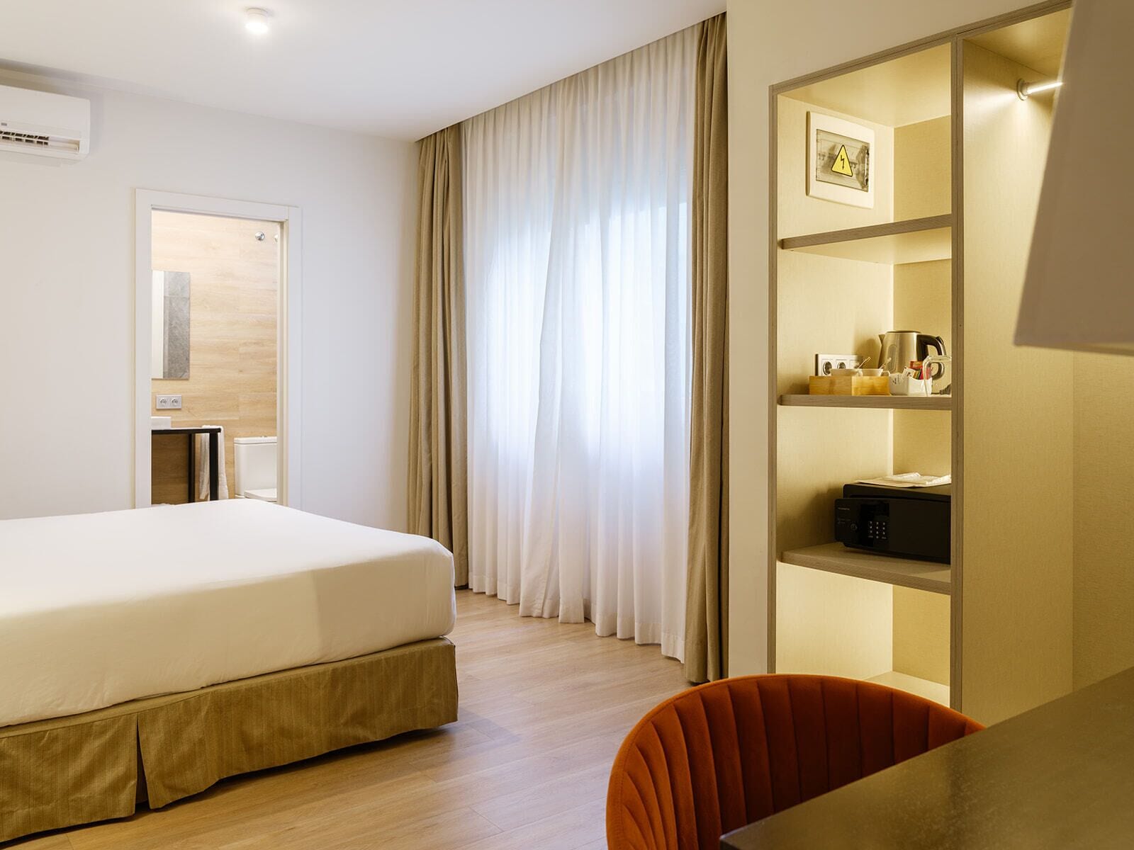 Une chambre d’hôtel comprenant un lit, un bureau, une chaise, une étagère avec quelques objets et une fenêtre avec des rideaux.