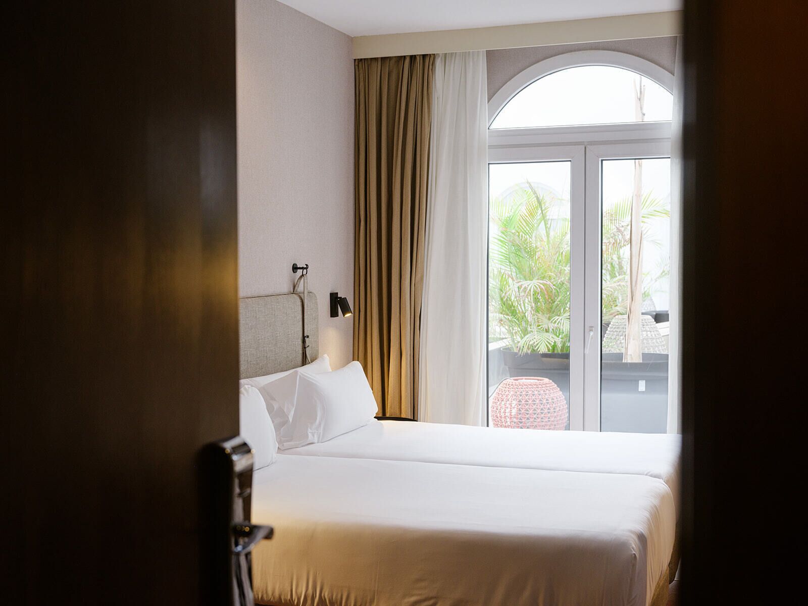 Une chambre d’hôtel avec un lit, une table de chevet, une lampe, une fenêtre avec des rideaux et une vue sur la verdure.