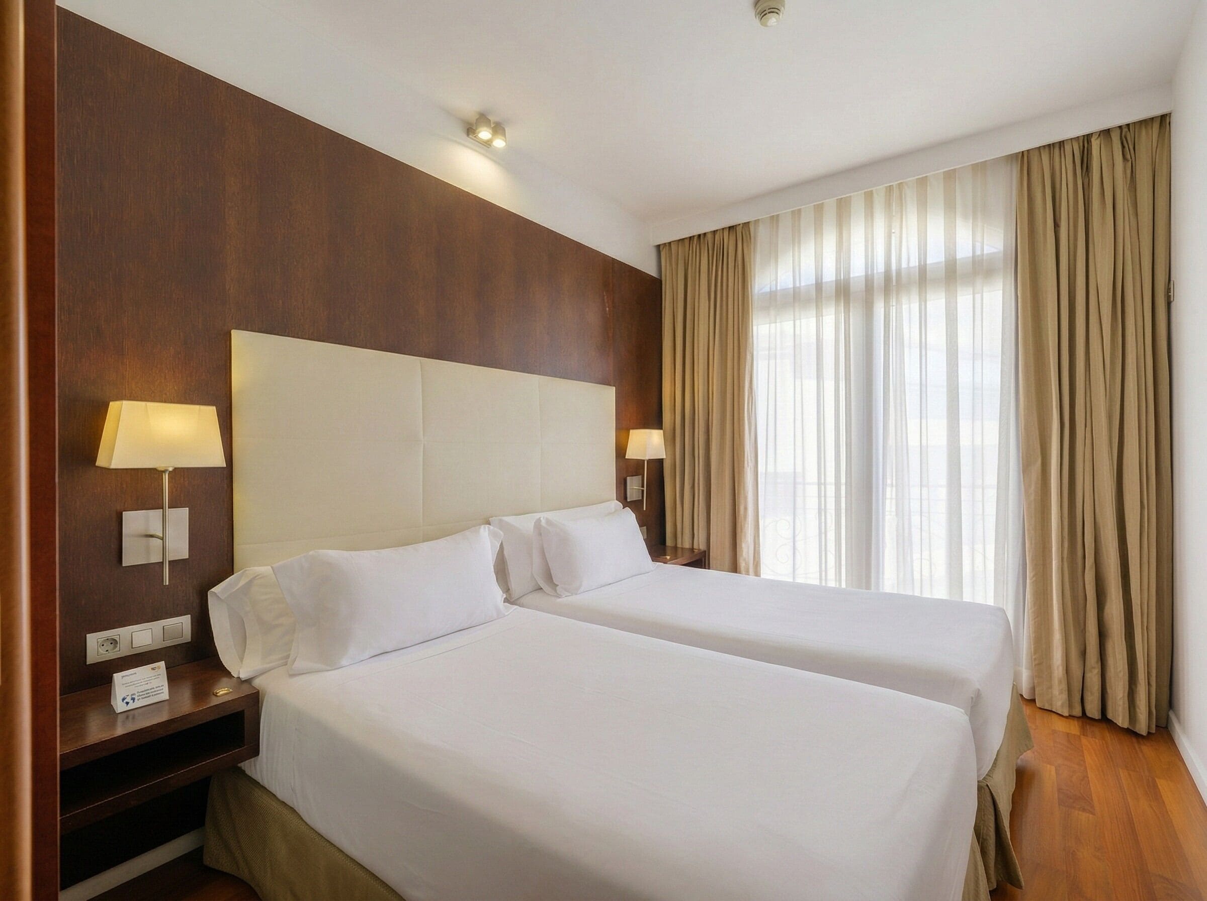 A hotel room with two beds, wooden headboard, bedside lamps, and a large window with curtains.