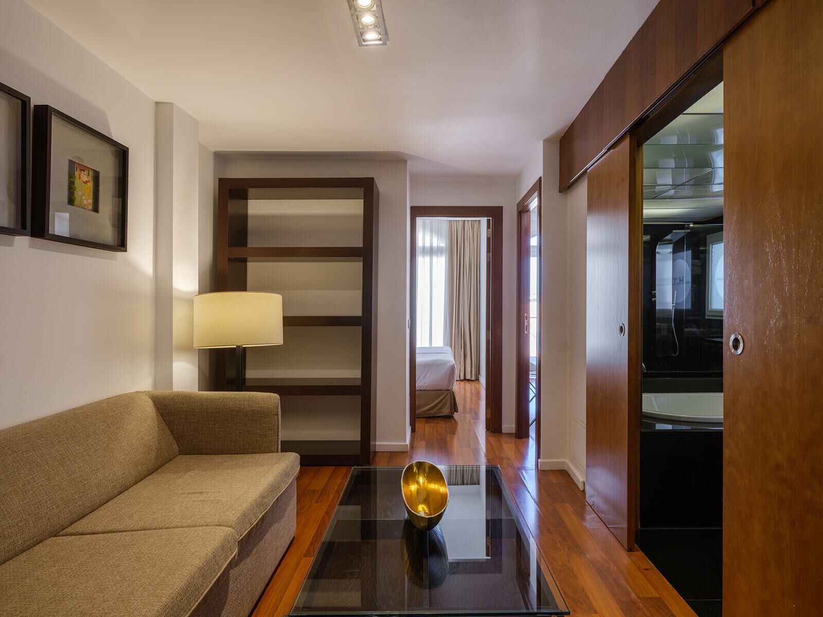 A hotel room with a sofa, a glass coffee table, a lamp, a wooden wardrobe, and a shower area.