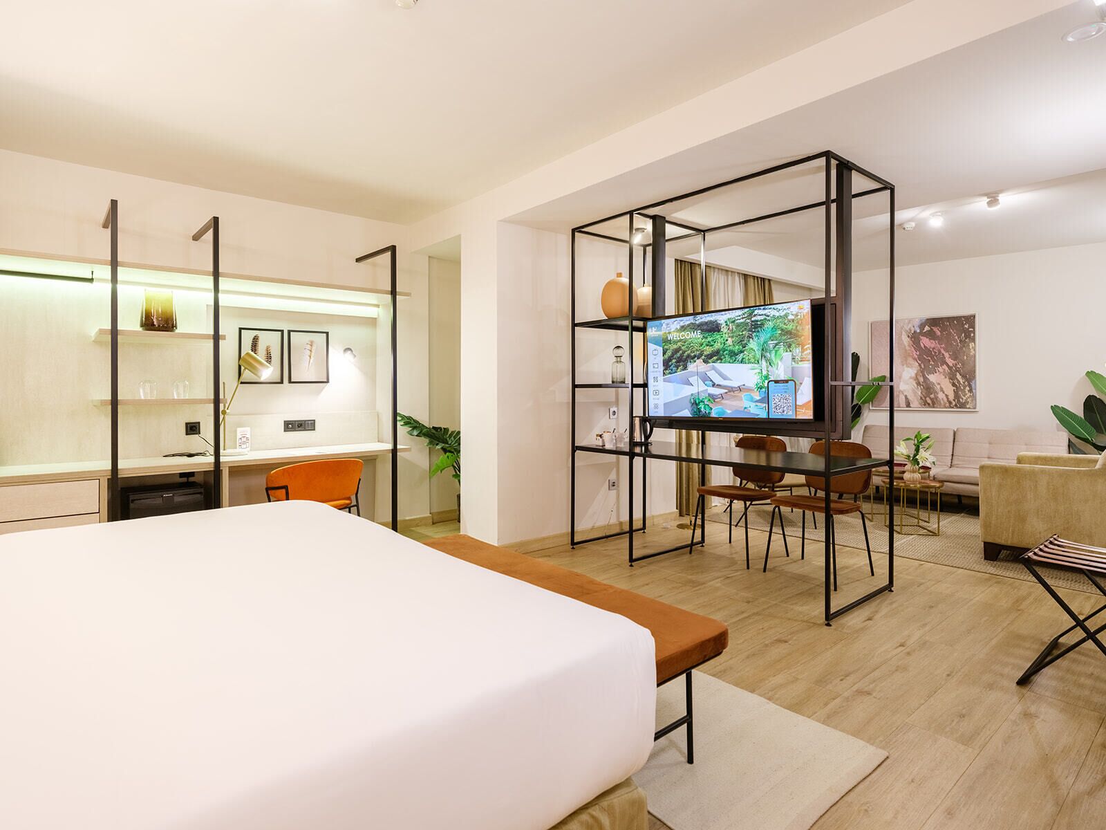 Una habitación de hotel moderna con una cama grande, un escritorio con una computadora, un televisor y una zona de estar con sofá y sillones.
