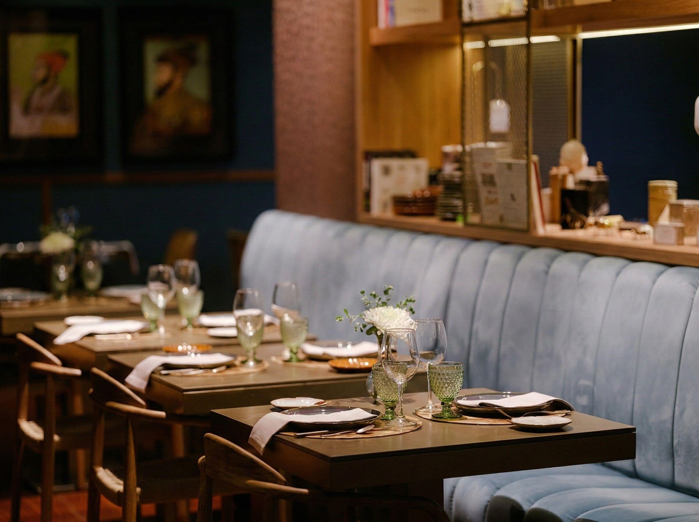 Un ristorante con un lungo tavolo di legno apparecchiato per la cena, una panca imbottita blu e uno scaffale con diversi oggetti sullo sfondo.