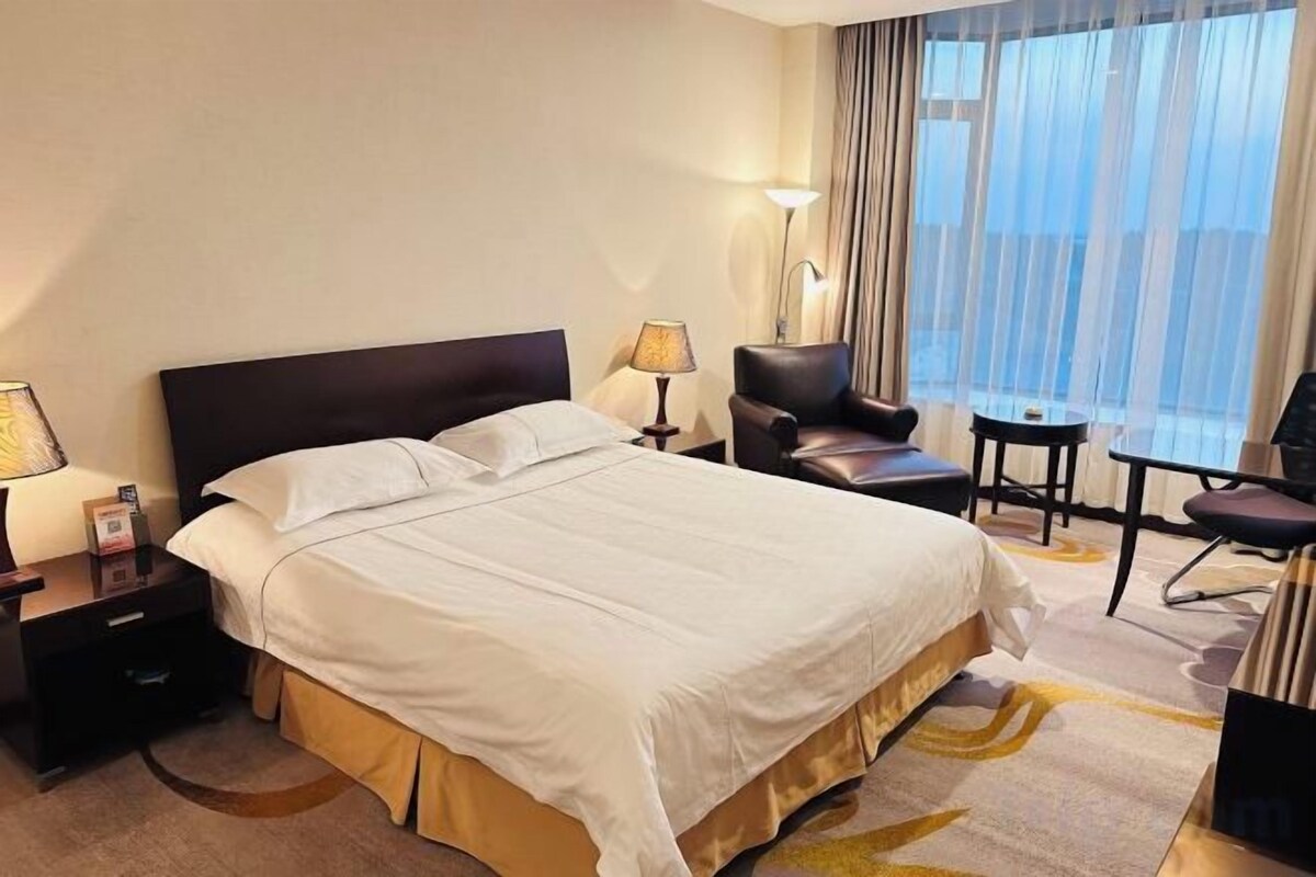 A hotel room with a large bed, bedside tables, a chair, a small table, and a view of the outdoors.