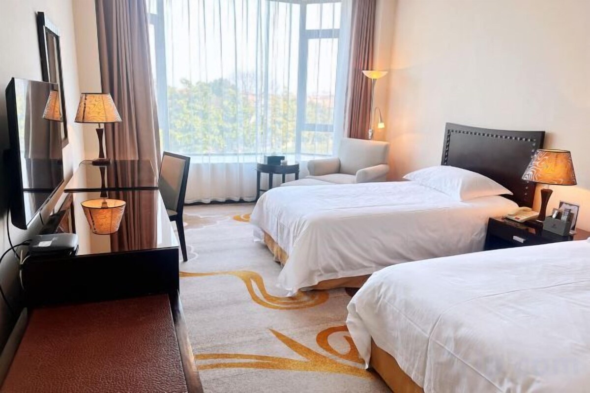 A hotel room with two beds, a desk with a lamp, a TV, and a view of the outdoors.