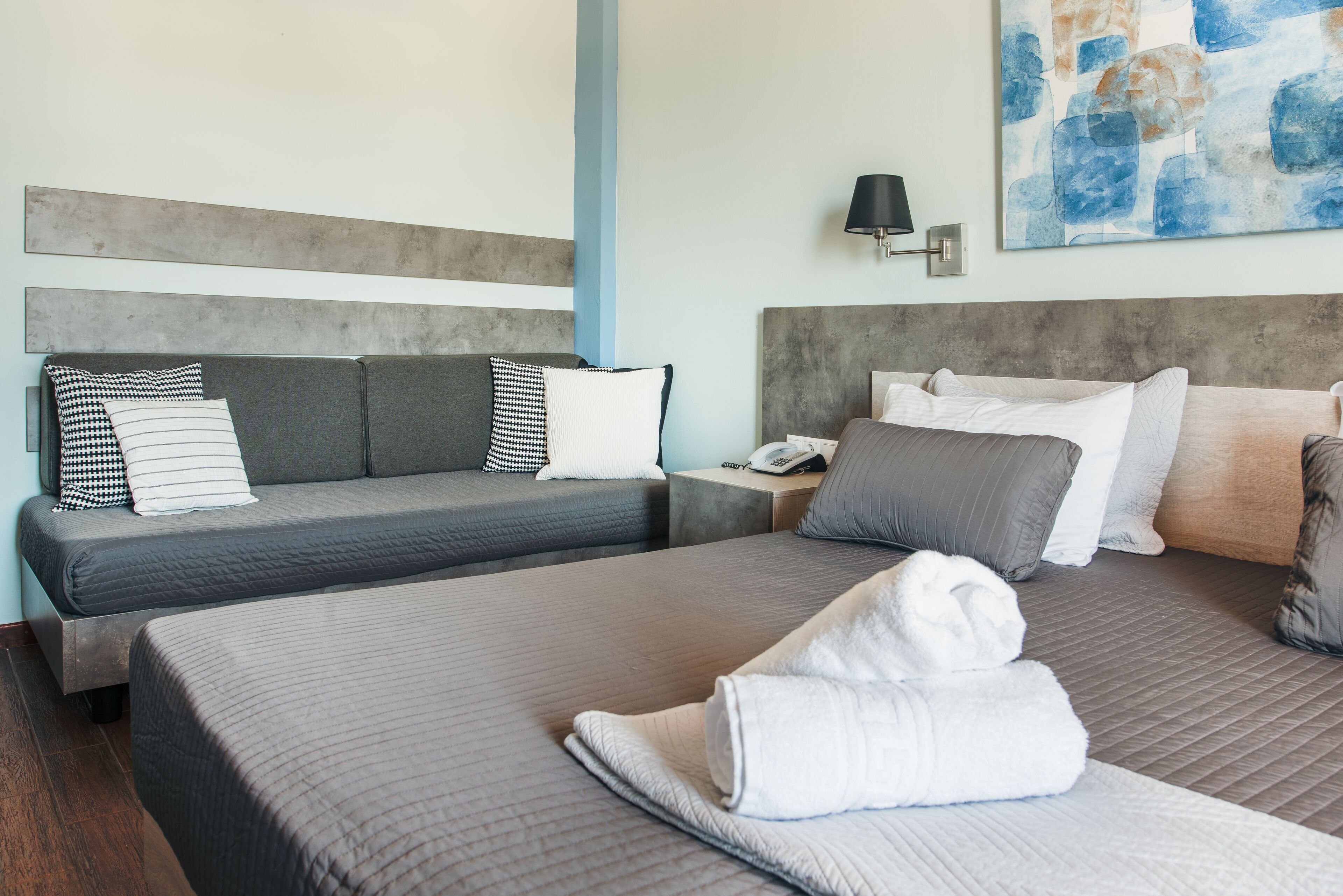 Quarto de hotel com duas camas, decoração em tons de cinza e branco, um quadro na parede e uma mesa de cabeceira com abajur.