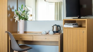 A hotel room with a wooden desk, a chair, a television, a vase with flowers, a kettle, and a shelf with towels. - Myrto Hotel (Marathon)