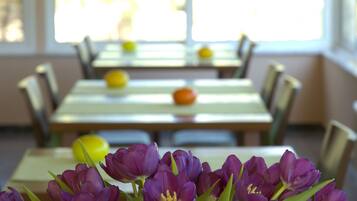 A dining area with tables and chairs, a vase of purple flowers in the foreground, and a bright window in the background.