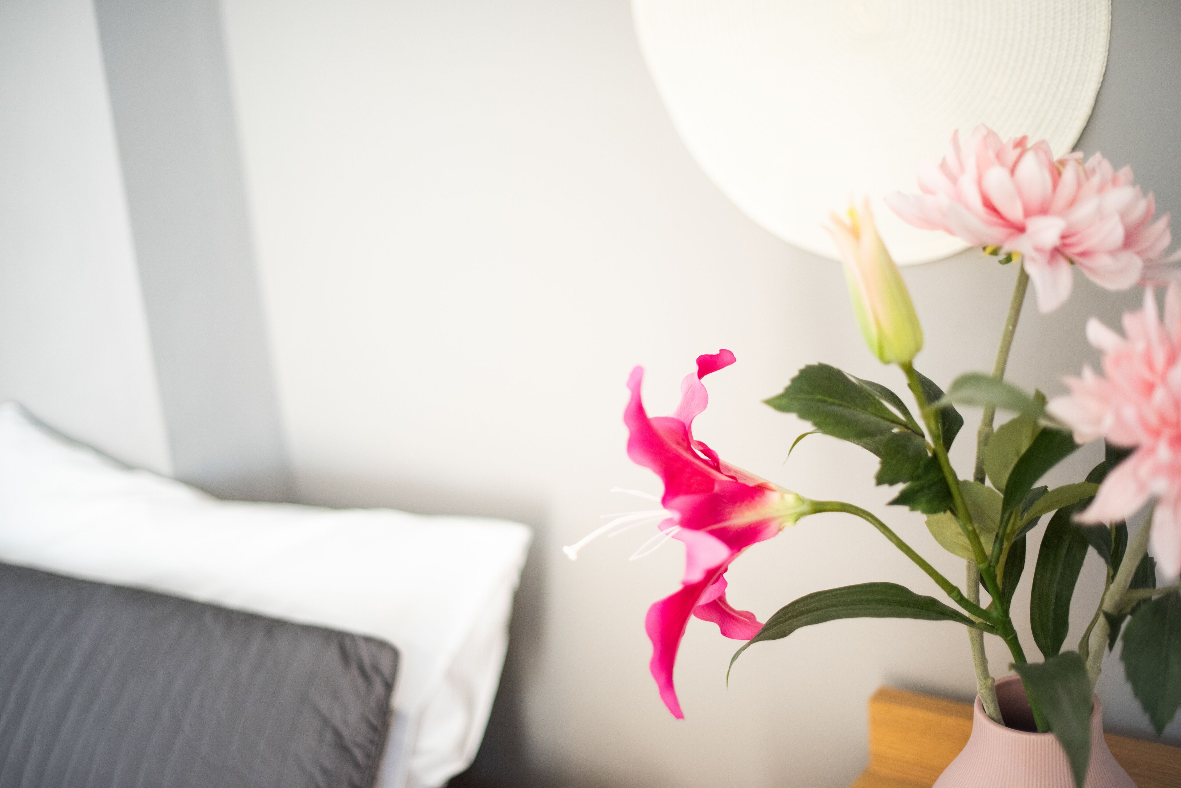 A bed with a white pillow and a bedside table with a vase of pink flowers.